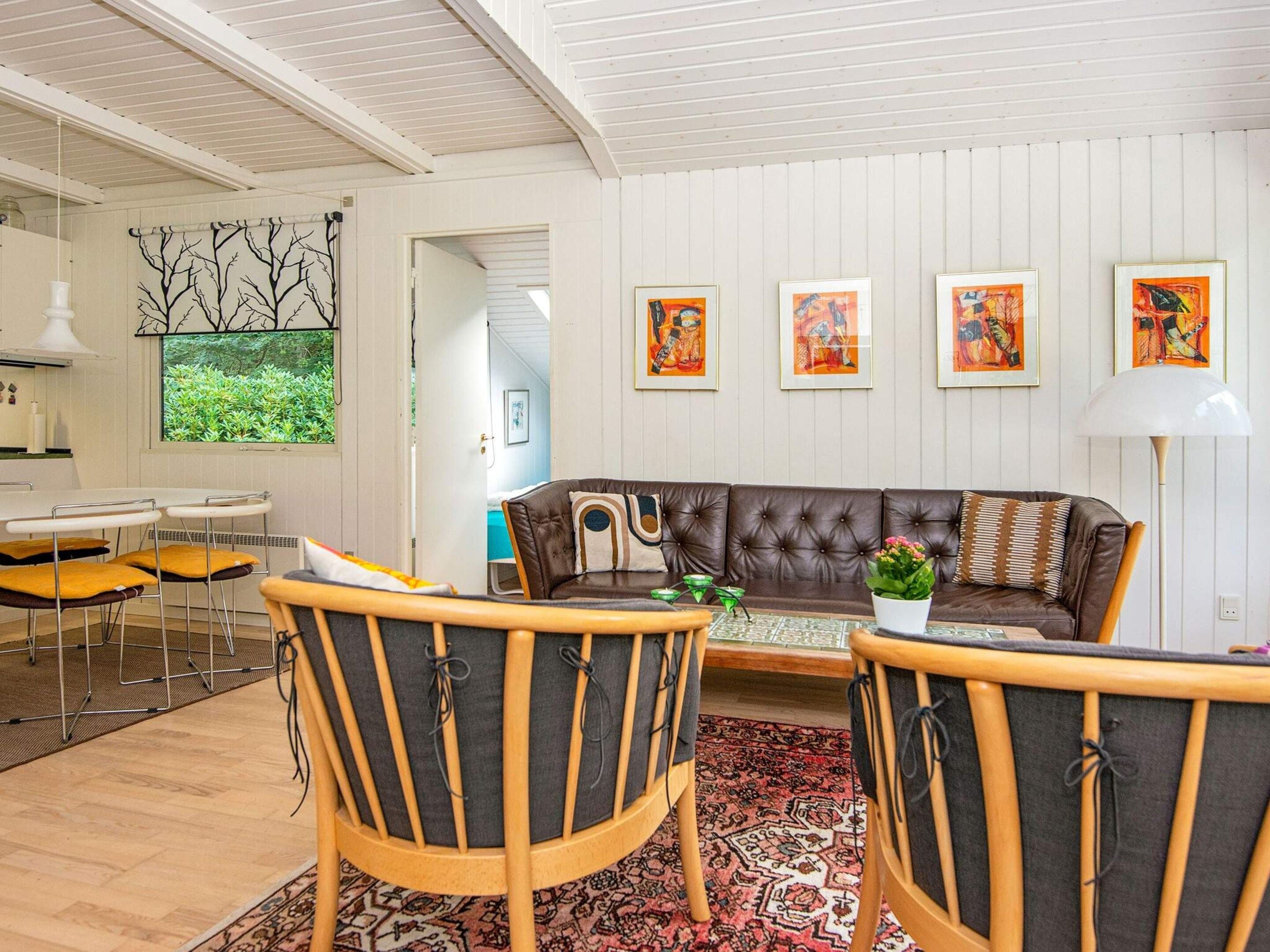 Una sala moderna con un sofá de cuero marrón, dos sillas de madera, una zona de comedor con una mesa y sillas blancas, y un ventanal con vista a zonas verdes.