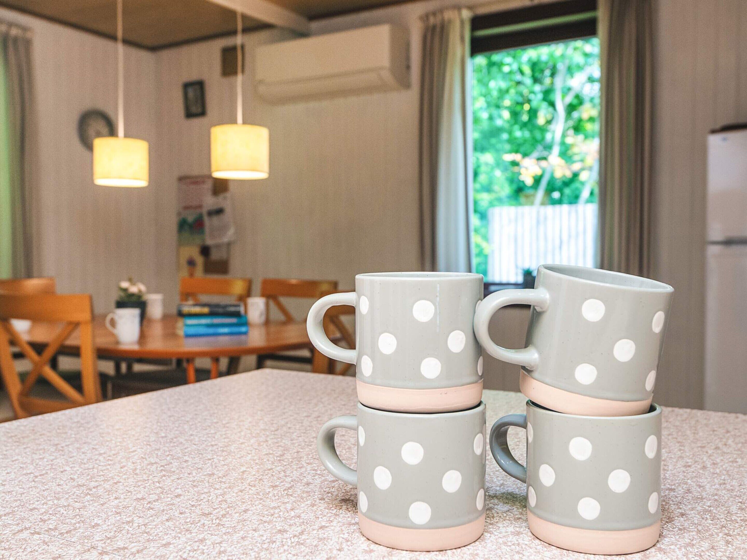 A dining area with a table and chairs, a window with a view of greenery, and a stack of polka-dotted mugs on a countertop.