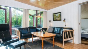 A modern living room with a wooden ceiling, a black leather sofa, a wooden coffee table, and a view of greenery outside.