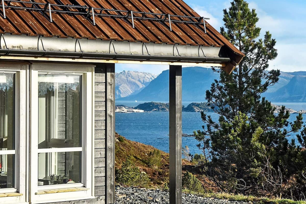 A rustic cabin with a wooden roof and large windows overlooking a coastal landscape with mountains in the background.