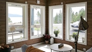 A cozy room with a grey sofa, a small round table, and a lamp. There are three windows with views of a lake and trees. - 6 Person Holiday Home in Kalvag (Bremanger)