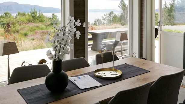 A dining area with a table set for two, a vase with white flowers, and a view of a lake and mountains through large windows. - 6 Person Holiday Home in Kalvag (Bremanger)
