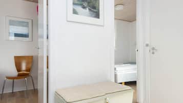 A room with a wooden floor, a white door, a bed with a white mattress, a small wooden chair, and a framed picture on the wall.