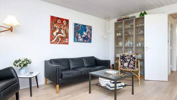 A modern living room with a black leather sofa, a glass coffee table, and a floral armchair. There are two abstract paintings on the wall, a lamp, and a glass cabinet with dishes.
