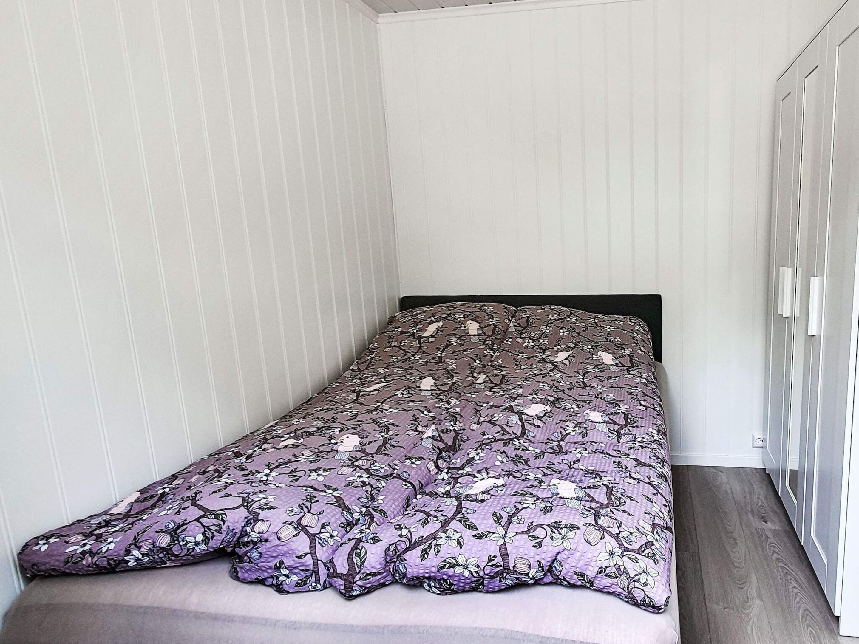 A small, single-bed room with a plain white wall and a wooden floor.