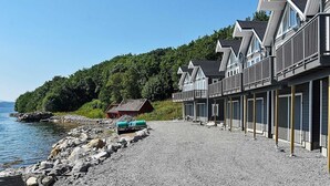 Een rij vakantiehuisjes aan het water, met balkons, een grindpad en een klein tuinhuisje bij de waterlijn.