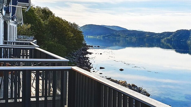 Een pand aan het water met een houten terras dat uitkijkt over een rustig meer, omgeven door bergen.