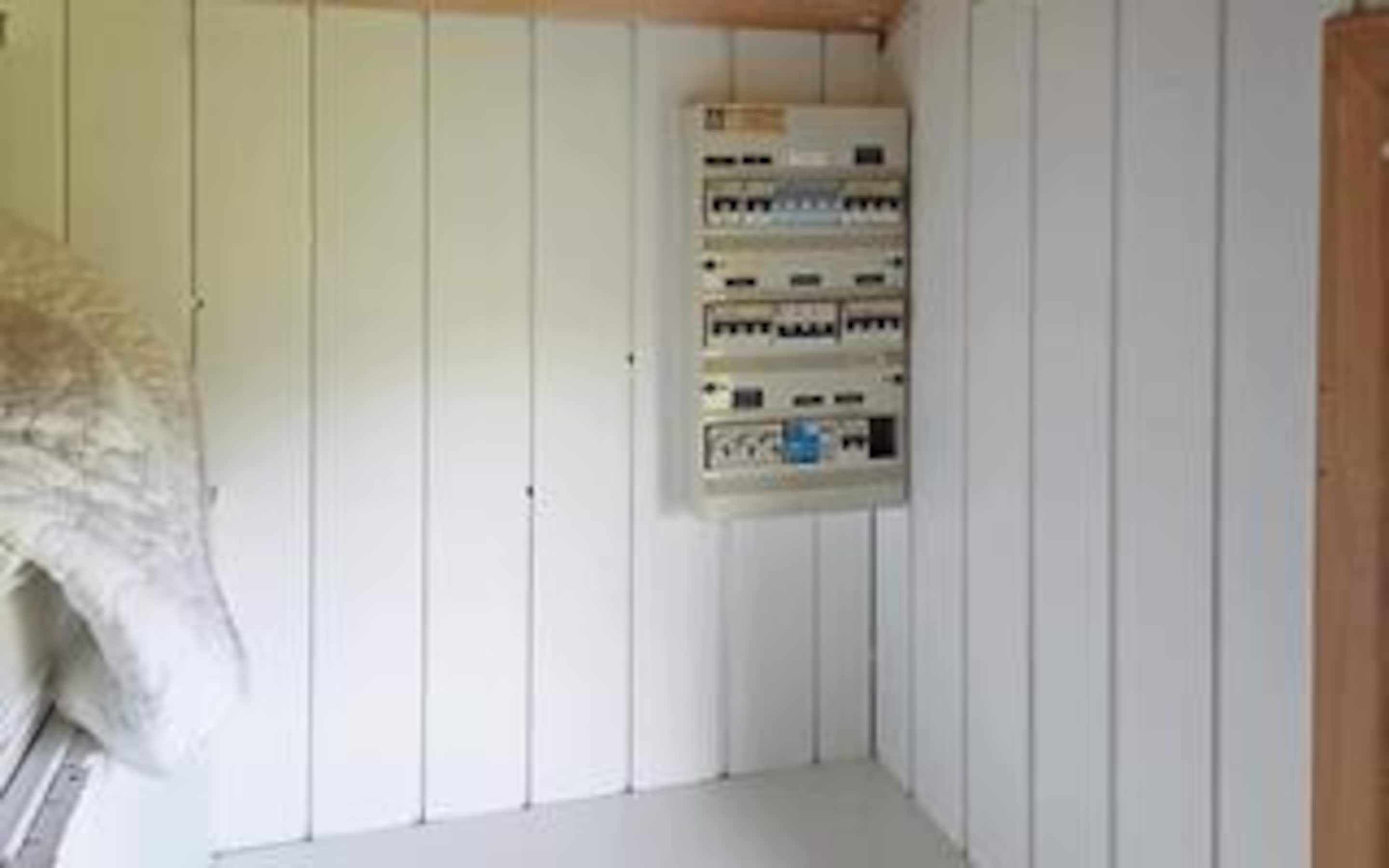 A room with white paneled walls, a white floor, and a wall-mounted electrical panel.