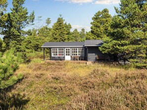A small cabin with a deck and outdoor seating, surrounded by dense trees and tall grass. - 4 Star Holiday Home in Romo (Rømø)