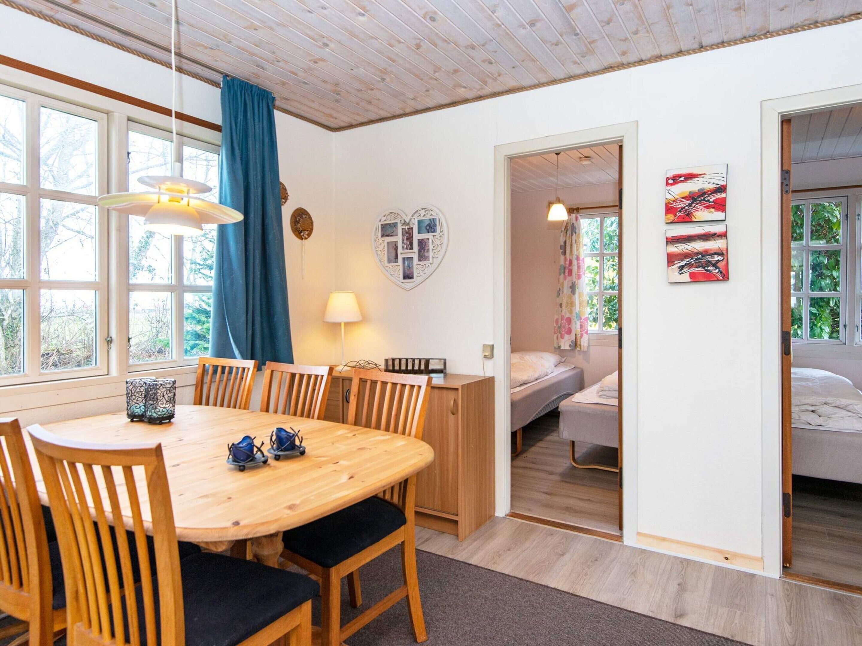 A room with a wooden dining table, chairs, a bed, and a window with curtains.