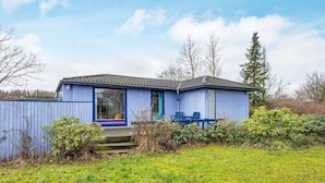 Une petite maison à un seul étage, avec une façade bleue et un toit recouvert de tuiles. Elle possède un porche à l’avant, avec deux chaises bleues et une petite table.