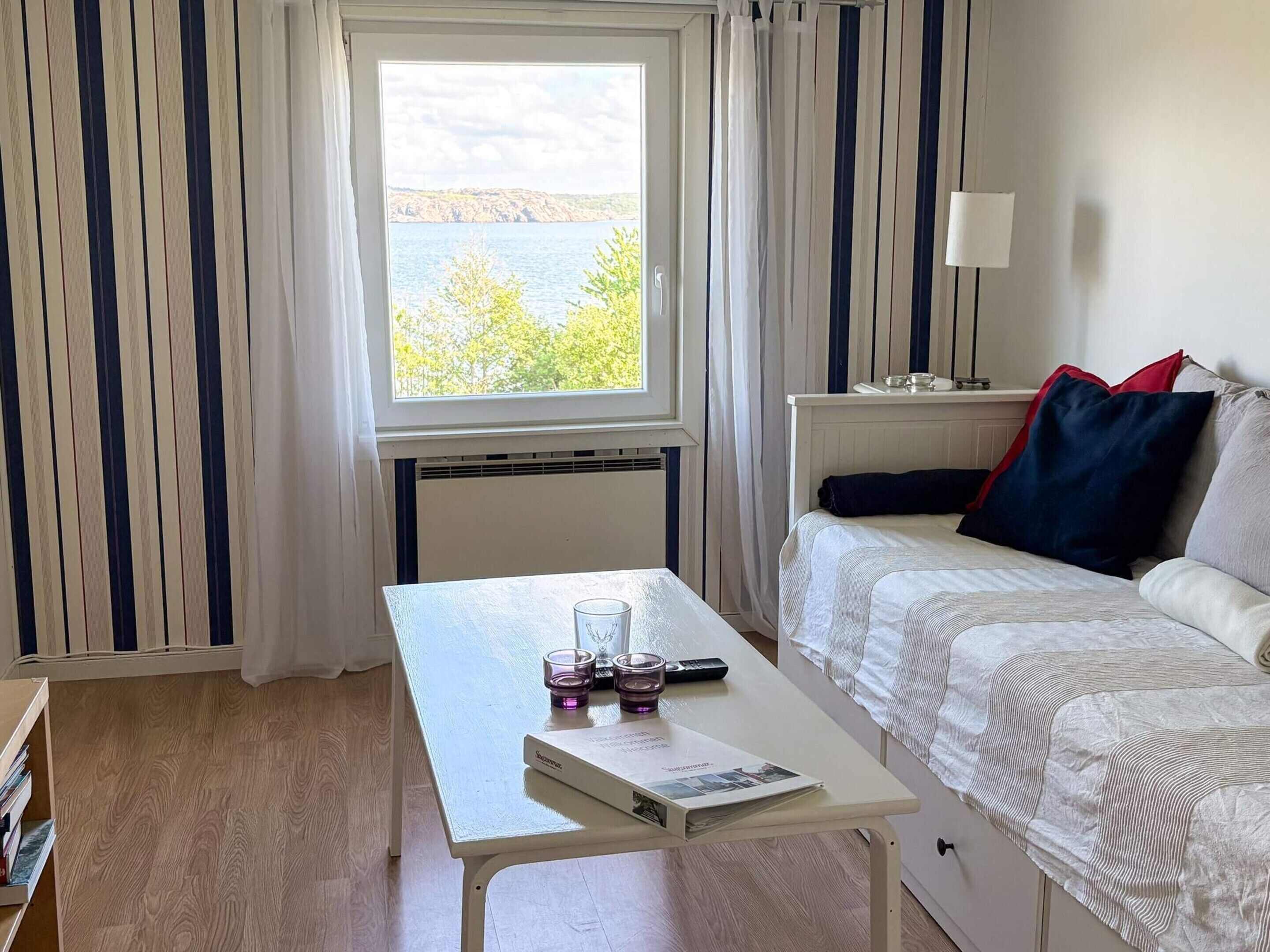 A room with a bed, a table with a book and glasses, a lamp, and a window with a view of trees and water.