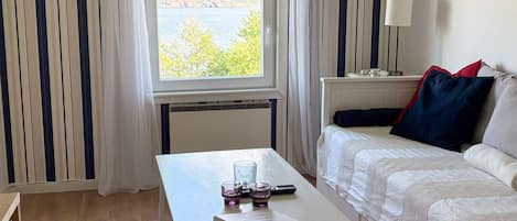 A room with a bed, a table with a book and glasses, a lamp, and a window with a view of trees and water.