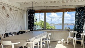 A dining area with a long table and white chairs, large windows with a view of the sea, and a patterned curtain. - 4 Star Holiday Home in Lysekil (Lysekil)