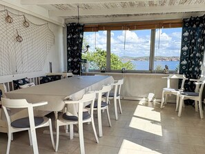 A dining area with a long table and white chairs, large windows with a view of the sea, and a patterned curtain.