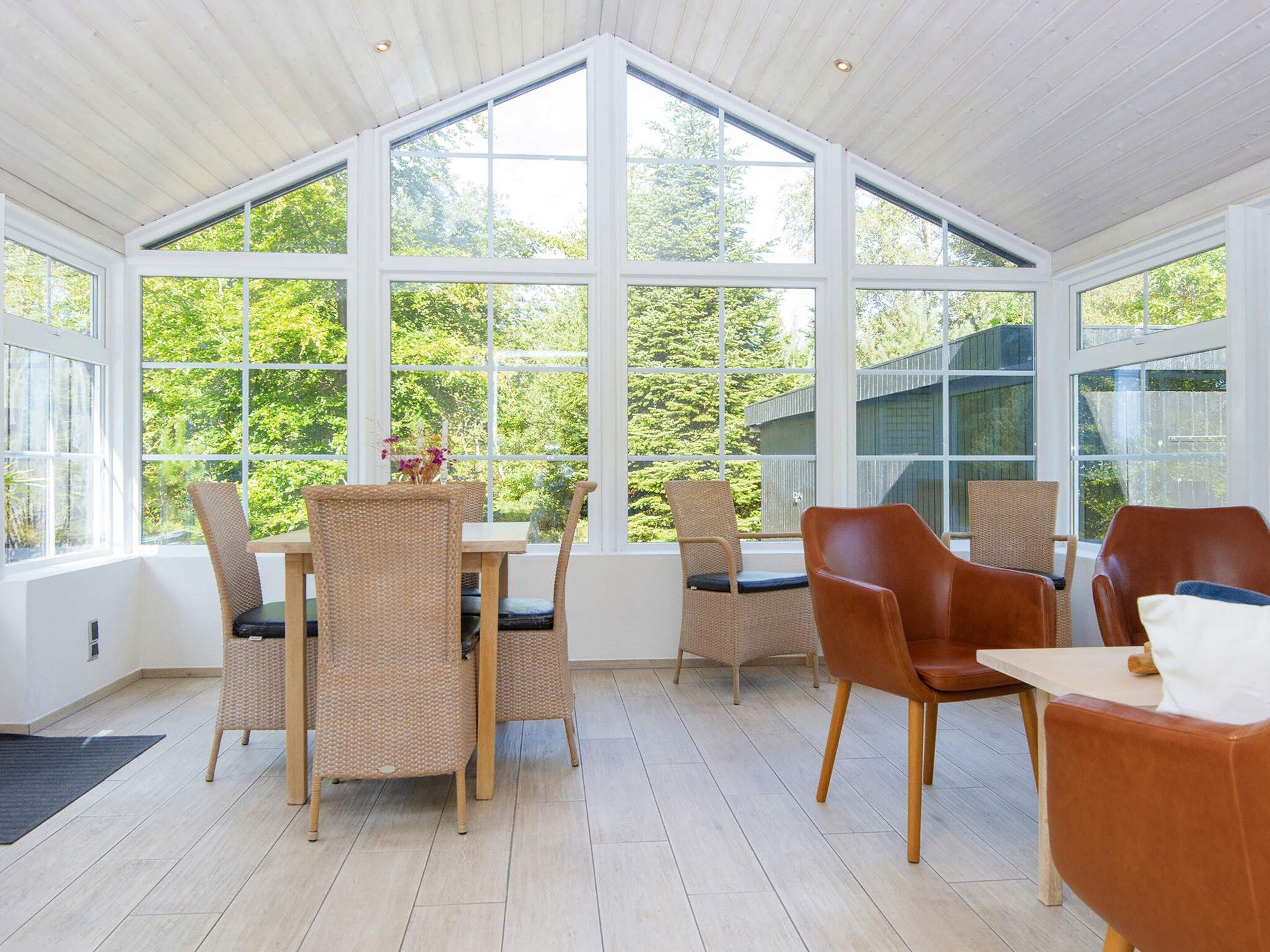 Une véranda dotée d’un haut plafond et de grandes fenêtres, aménagée avec des fauteuils en osier et une table en bois, et offrant une vue sur un paysage verdoyant.