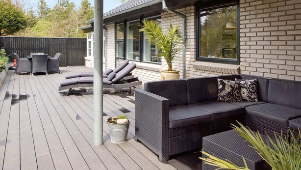 Eine überdachte Terrasse im Freien mit einer dunkelgrauen Couch, einem Esstisch mit Stühlen und einem Blumentopf.
