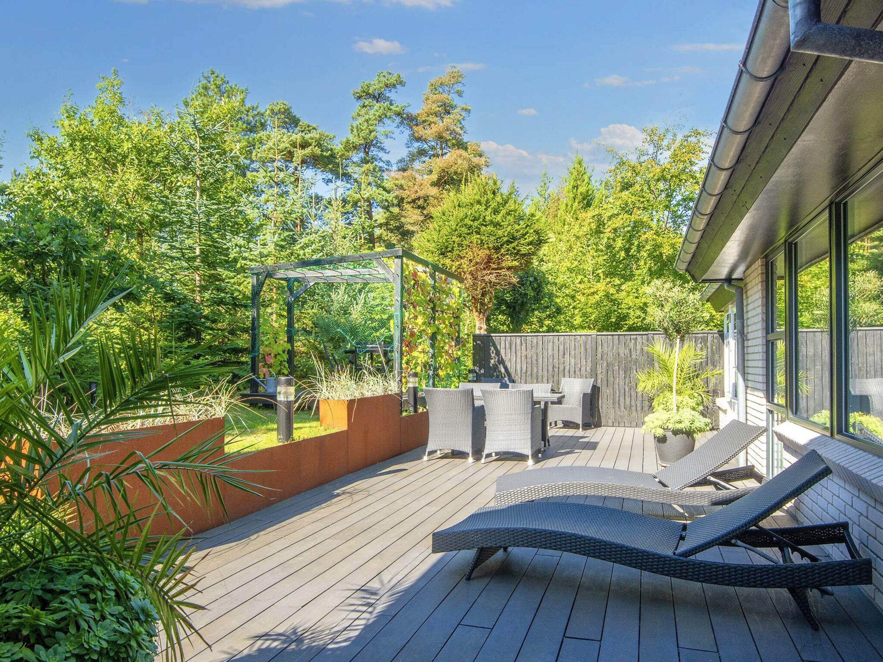 A deck with outdoor furniture, a pergola, and a view of a garden and trees.