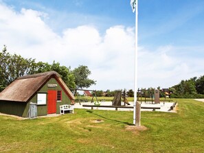 A rustic building with a thatched roof and a red door, a playground with various equipment, and a flagpole in a grassy area. - 4 Person Holiday Home in Hemmet (Hemmet)