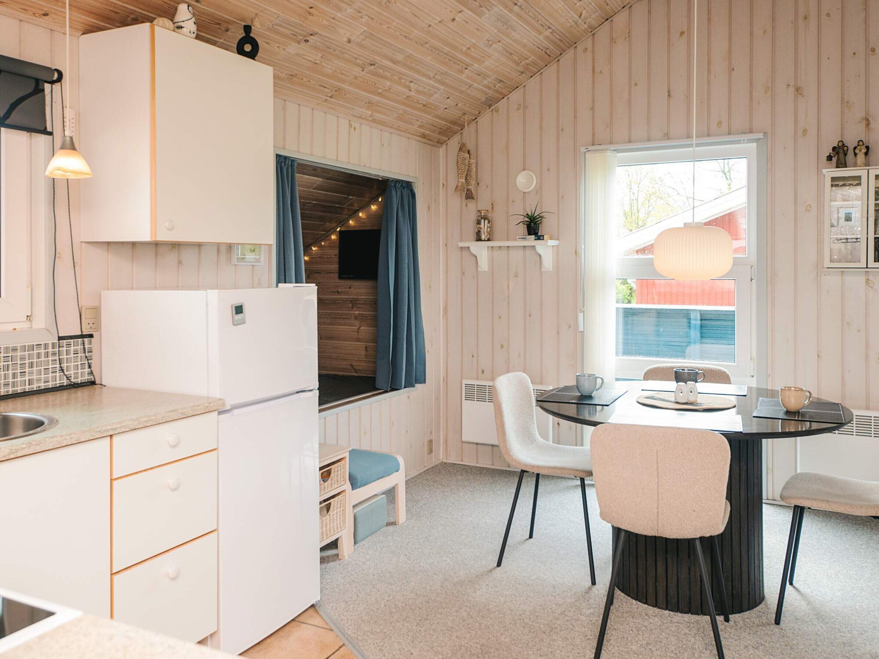 A compact kitchen with a refrigerator, a dining table with chairs, a window with curtains, and a small shelf with decorative items.