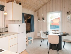 A compact kitchen with a refrigerator, a dining table with chairs, a window with curtains, and a small shelf with decorative items. - 4 Person Holiday Home in Hemmet (Hemmet)