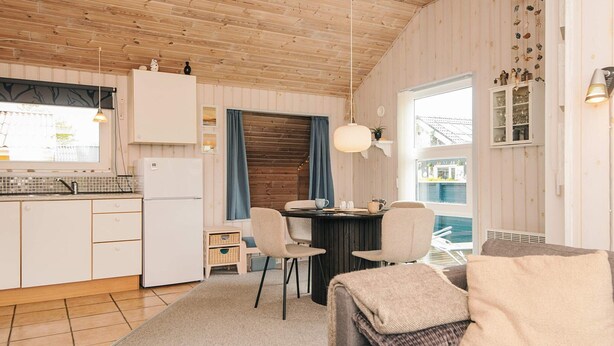 A cozy living room with a sofa, a dining table with chairs, a kitchen area with a refrigerator, and a view of a pool through a window. - 4 Person Holiday Home in Hemmet (Hemmet)