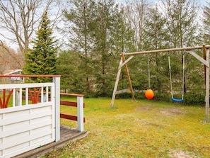A wooden swing set with two swings, one orange and one blue, in a grassy yard with trees in the background.