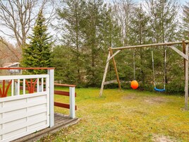A wooden swing set with two swings, one orange and one blue, in a grassy yard with trees in the background.