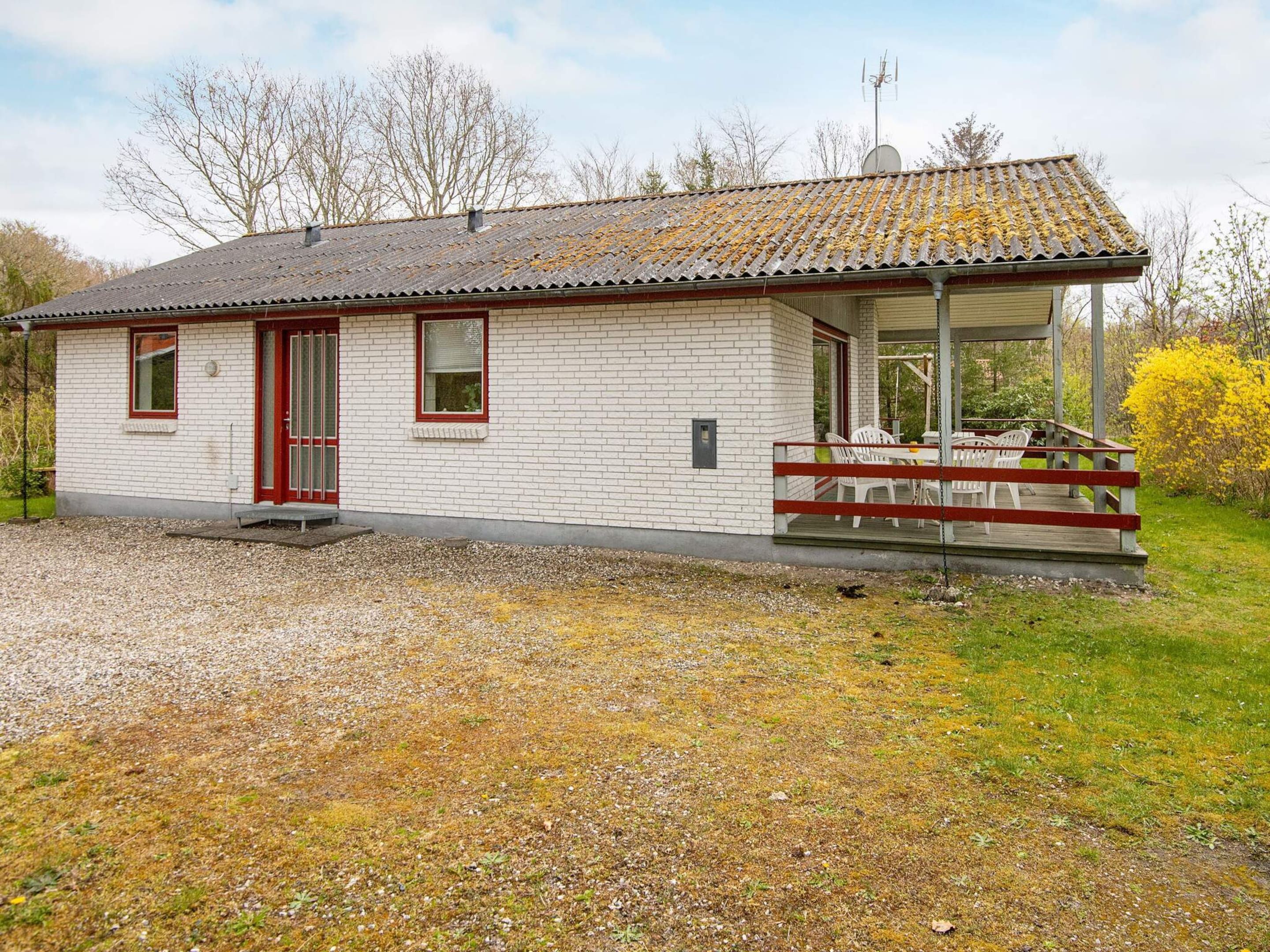 Une petite maison de plain-pied avec une porte rouge et des murs blancs, un porche couvert avec des chaises blanches et une allée gravillonnée.