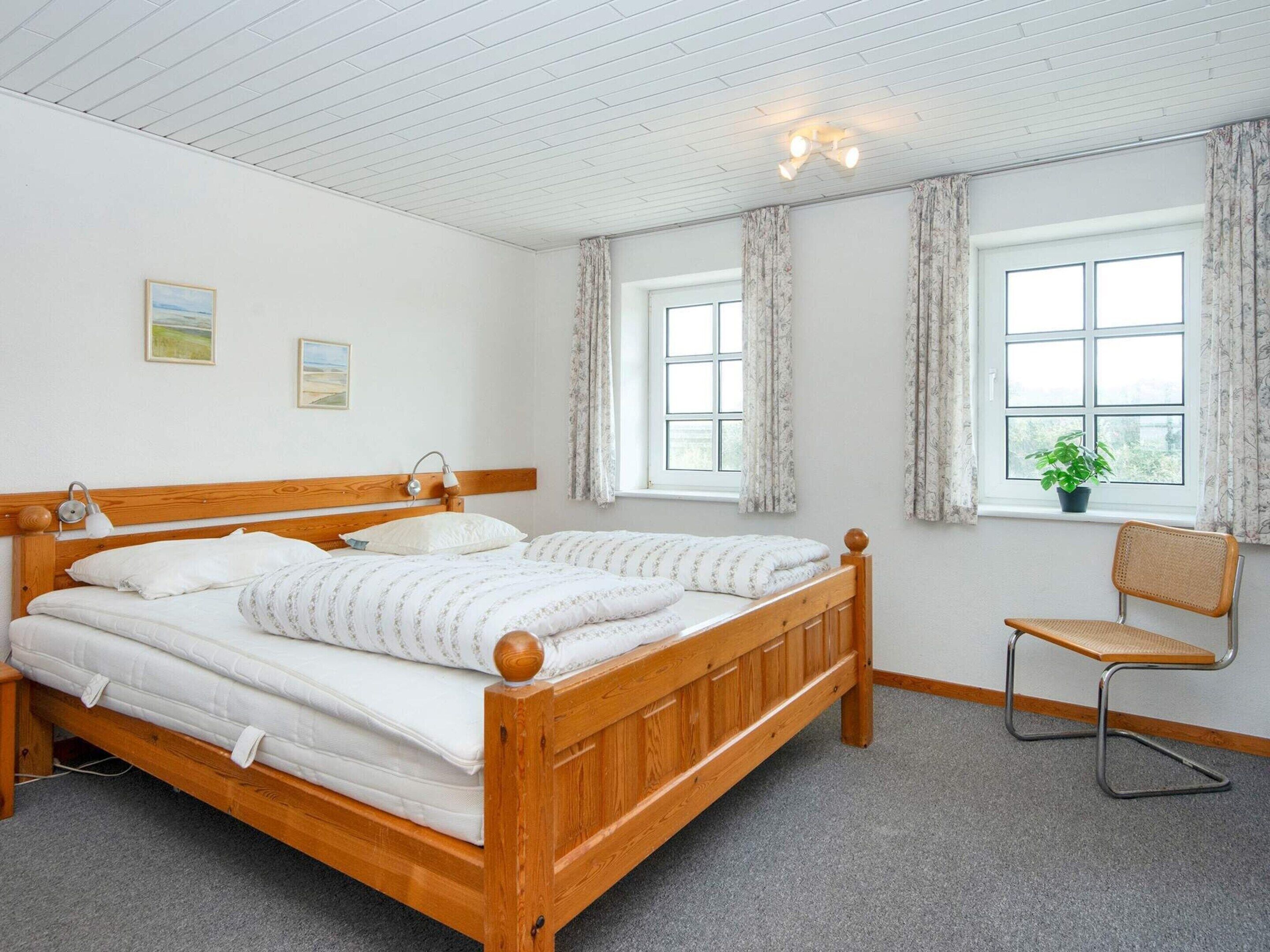A double bed with a wooden headboard, two bedside lamps, and a small chair. There are two windows with curtains, a potted plant on the windowsill, and a carpeted floor.