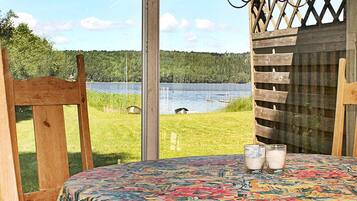 Una habitación con un amplio ventanal con vistas a un lago, una silla de madera, una mesa con mantel colorido y dos vasos de leche.