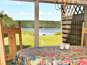 Una habitación con un amplio ventanal con vistas a un lago, una silla de madera, una mesa con mantel colorido y dos vasos de leche.