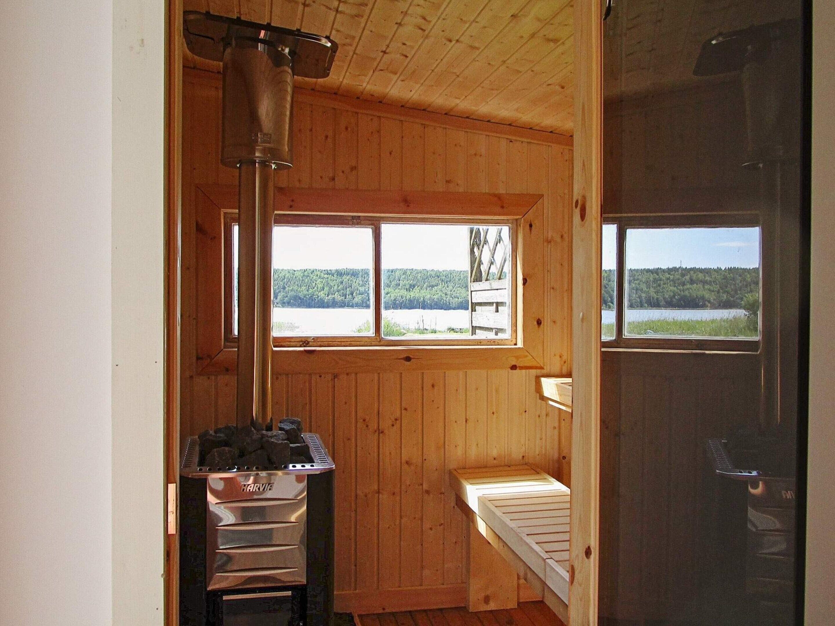 Interior de una cabaña de madera con un calentador de metal, un banco y ventanas con vistas a un paisaje boscoso.
