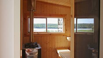 Interior de una cabaña de madera con un calentador de metal, un banco y ventanas con vistas a un paisaje boscoso.