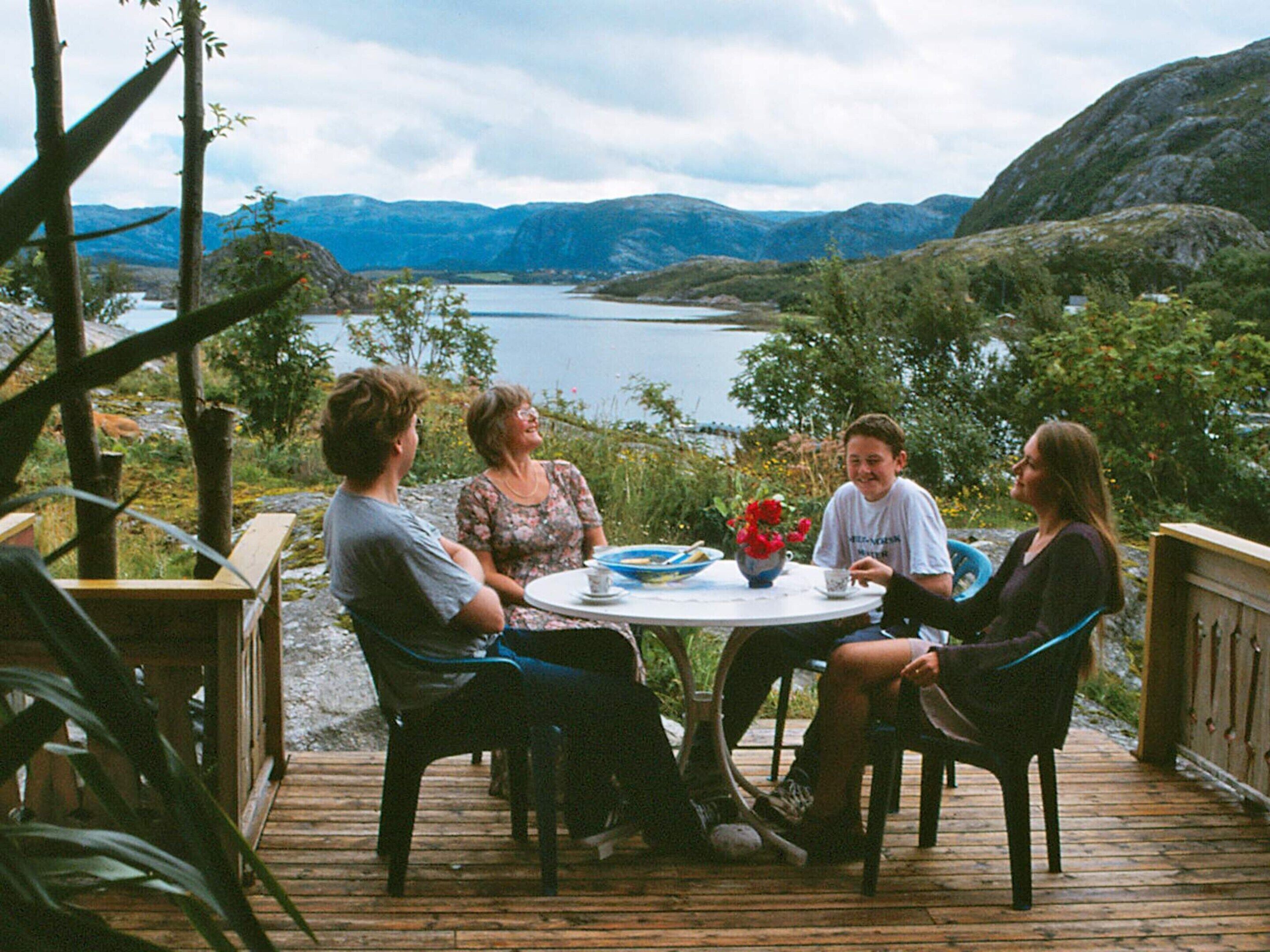 A group of people seated at a round table on a wooden deck with a lake and mountains in the background.