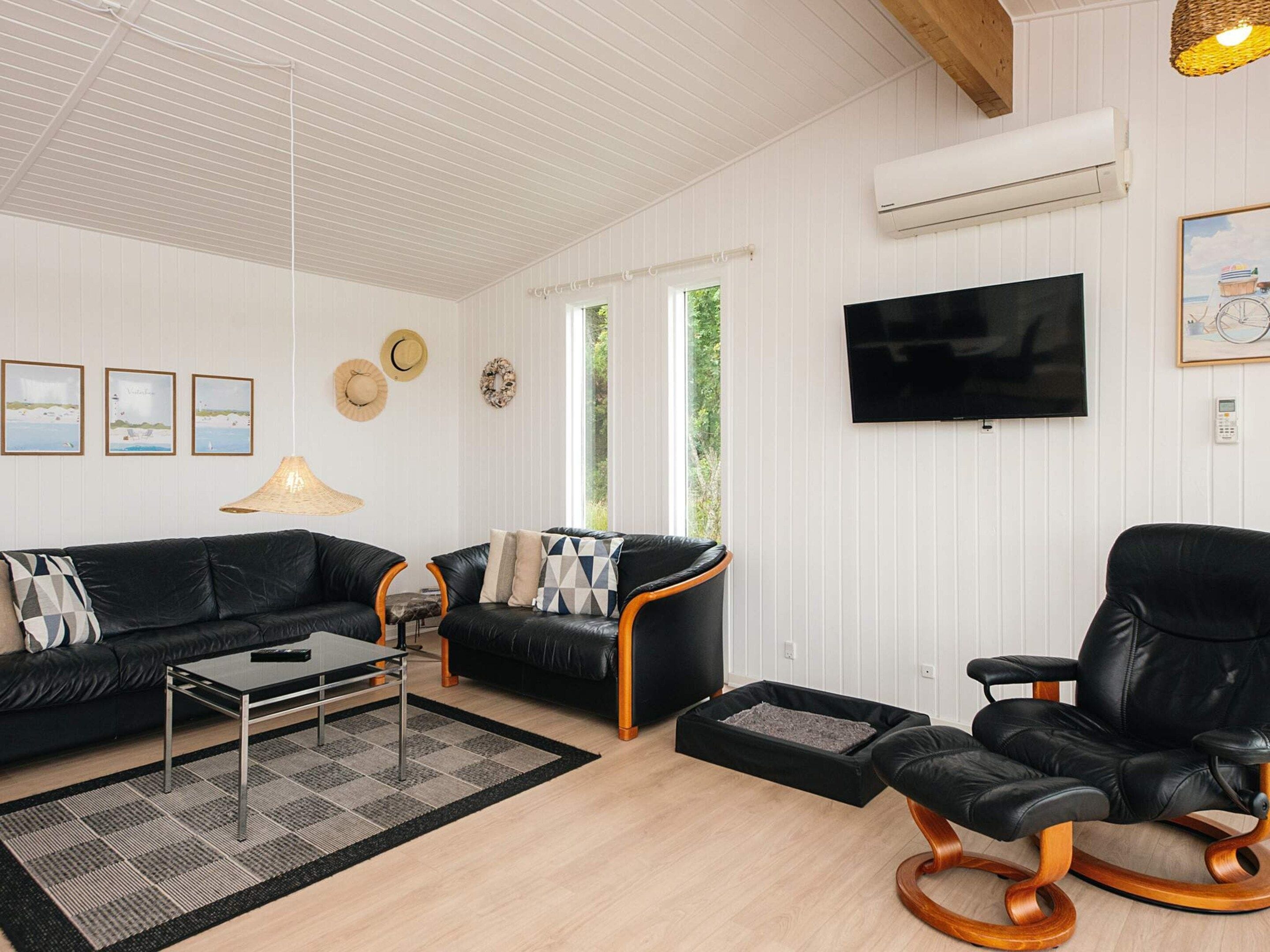 A living room with a black leather sofa, a coffee table, a black leather armchair, a TV mounted on the wall, and a window with a view of greenery.