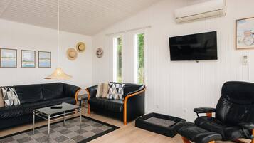 A living room with a black leather sofa, a coffee table, a black leather armchair, a TV mounted on the wall, and a window with a view of greenery.