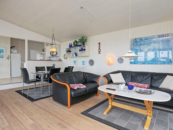A modern living room with a black leather sofa, a round coffee table, and a dining area with a white refrigerator and a hanging light fixture.