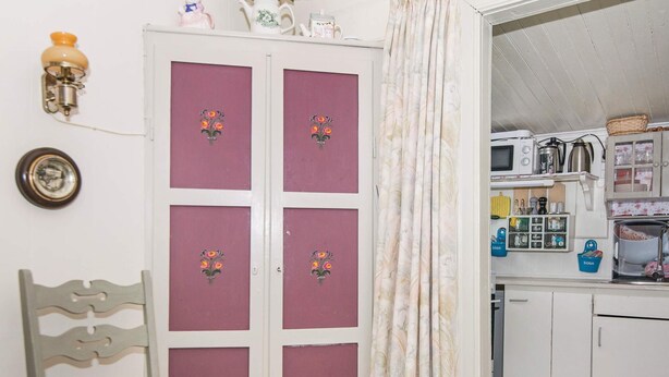 A room with a bed, a chair, a wardrobe with floral decorations, a kitchen area with appliances, and a checkered floor.