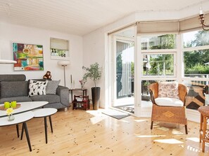 A modern living room with a grey sofa, a white coffee table, wicker chairs, and a large window with a view of greenery.