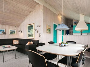 Una habitación acogedora con techo de madera, una mesa de comedor blanca con sillas, un sofá negro con cojines y una zona de cocina con armarios color turquesa.