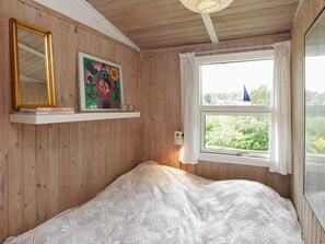 A bedroom with wooden walls, a bed with a white knitted blanket, a shelf with a framed picture and a book, and a window with a view of greenery. - 4 Person Holiday Home in Hojslev (Hojslev)