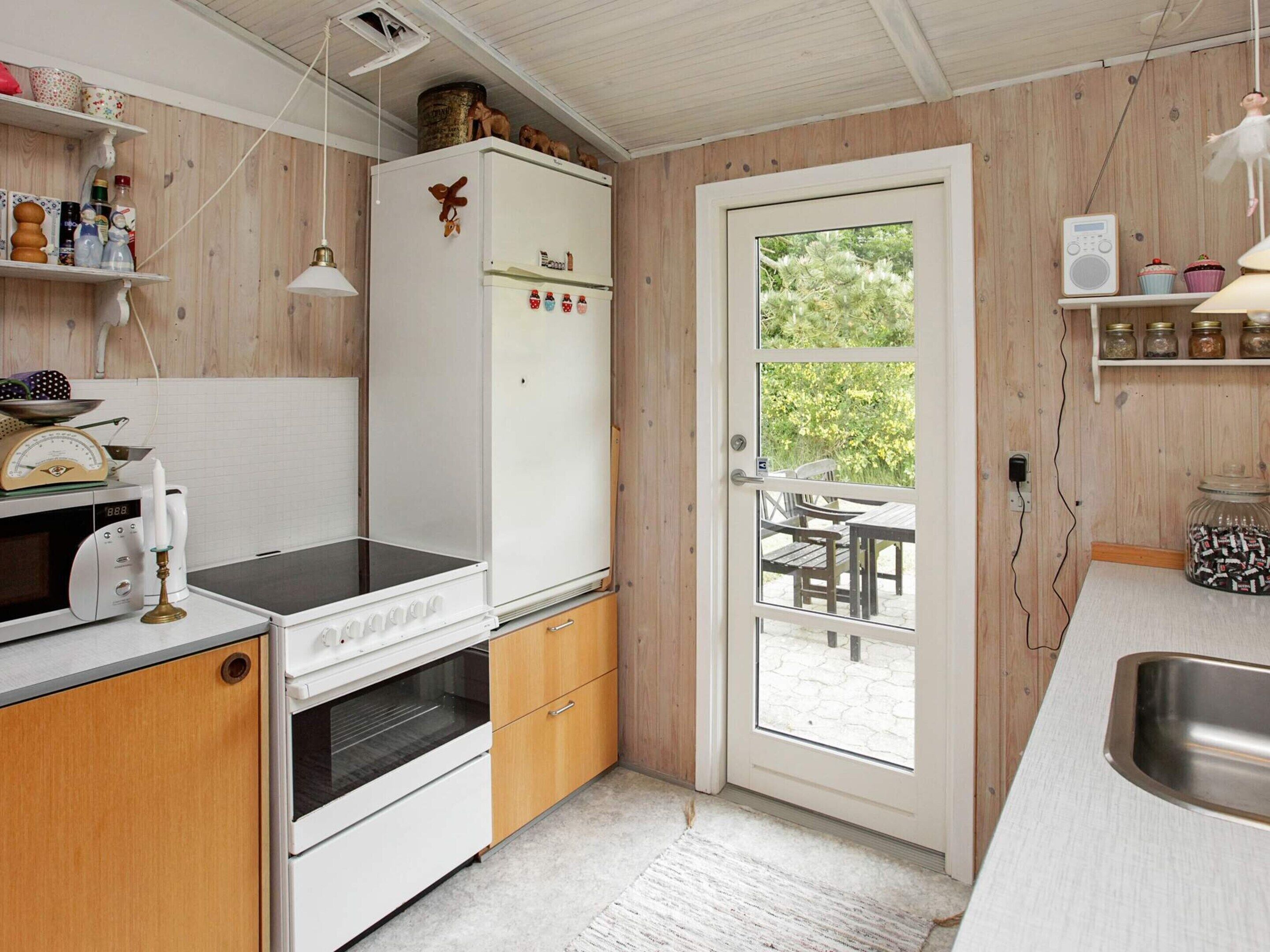 A compact kitchen with a microwave, oven, and refrigerator, a wooden door leading to an outdoor seating area, and a radio on the wall.