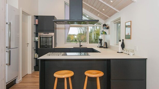 A modern kitchen with a large island, two wooden stools, a built-in oven, and a window overlooking a garden. - Splendid Holiday Home in Jutland With Whirlpool (Sydals)