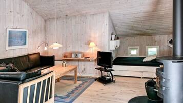 A cozy cabin interior with a wooden floor, a black leather sofa, a wooden table, a TV on a cart, a bed with a green mattress, and a wood-burning stove.