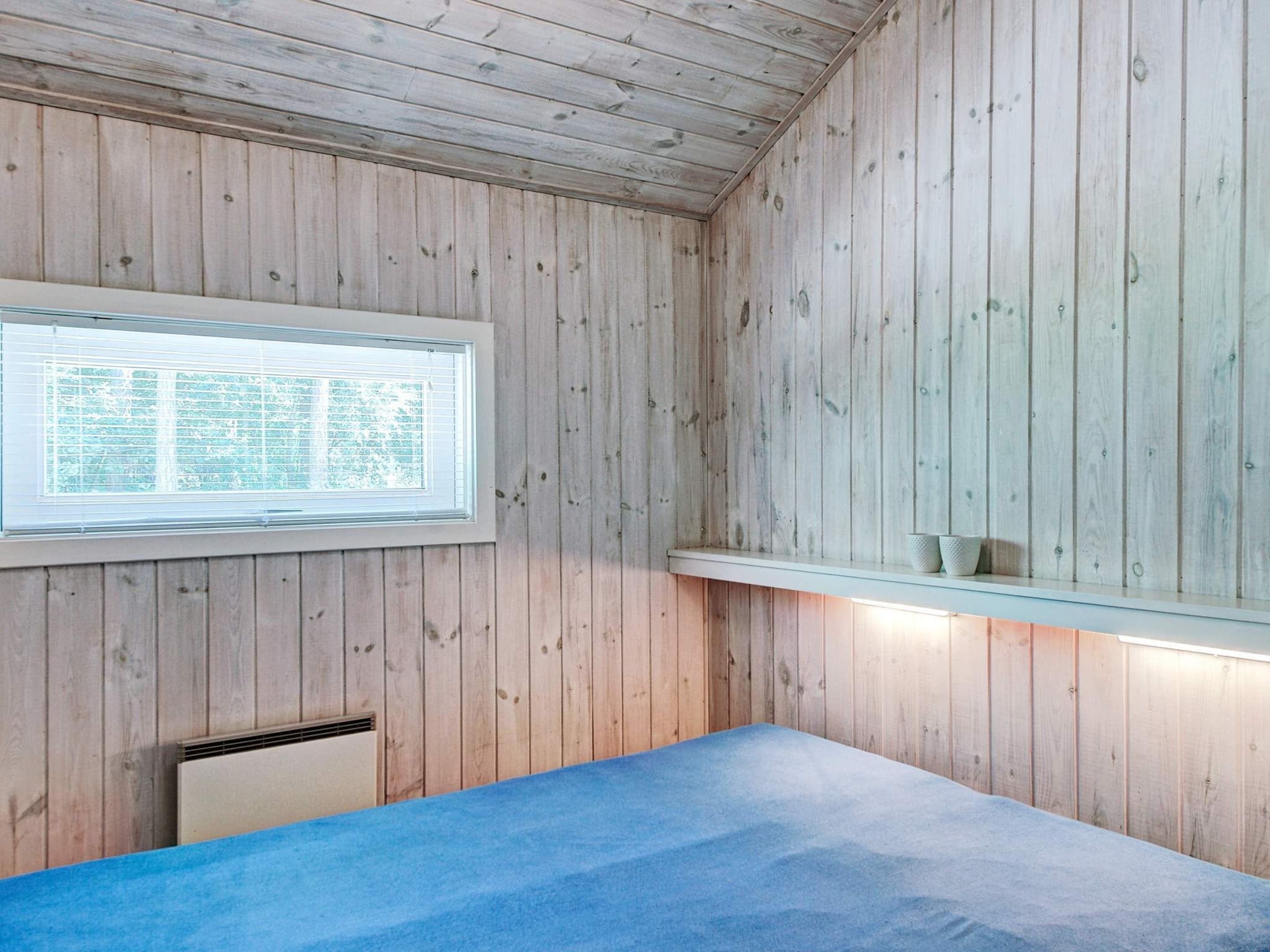 Quarto com paredes de madeira, uma cama com colchão azul, uma janela com persianas e uma prateleira com dois objetos.
