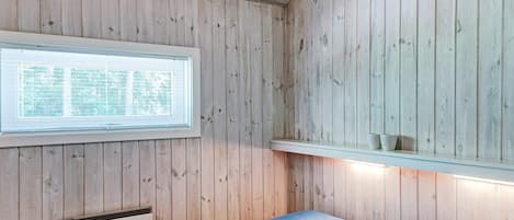 A room with wooden walls, a bed with a blue mattress, a window with blinds, and a shelf with two items.