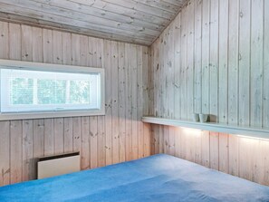 A room with wooden walls, a bed with a blue mattress, a window with blinds, and a shelf with two items. - Quaint Holiday Home in NexÃ¸ near Beach (Nexø)