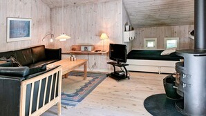 Interior acolhedor de uma cabana com teto de madeira, sofá de couro preto, mesa de jantar de madeira, televisão sobre um carrinho, lareira e uma cama com vista para o exterior.