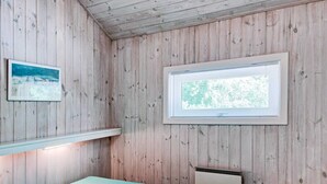 Quarto com paredes de madeira, uma cama com colcha azul clara, uma janela com vista para árvores e um quadro na parede.